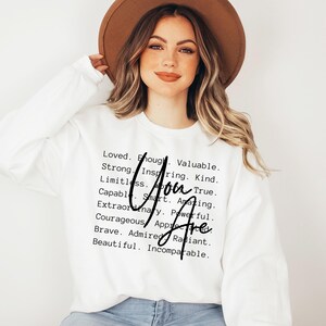 May include: White sweatshirt with black text that reads "You Are" and a list of positive attributes, including loved, enough, valuable, strong, inspiring, kind, limitless, true, capable, smart, amazing, extraordinary, powerful, courageous, appreciated, brave, admired, radiant, beautiful, and incomparable.