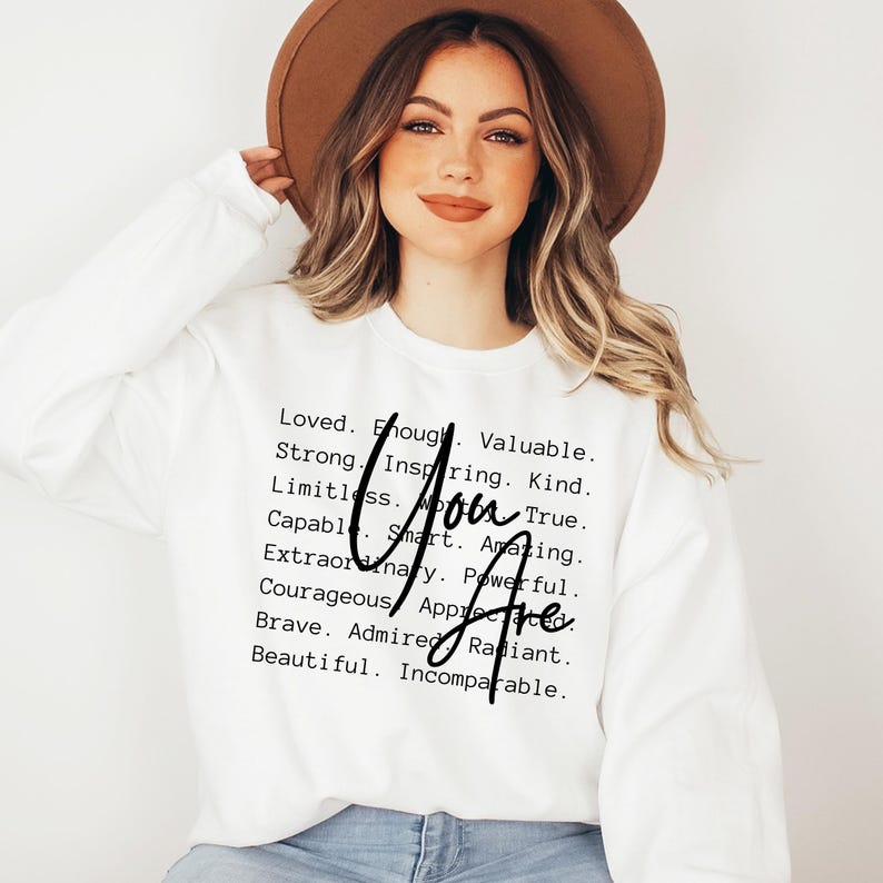 May include: White sweatshirt with black text that reads "You Are" and a list of positive attributes, including loved, enough, valuable, strong, inspiring, kind, limitless, true, capable, smart, amazing, extraordinary, powerful, courageous, appreciated, brave, admired, radiant, beautiful, and incomparable.