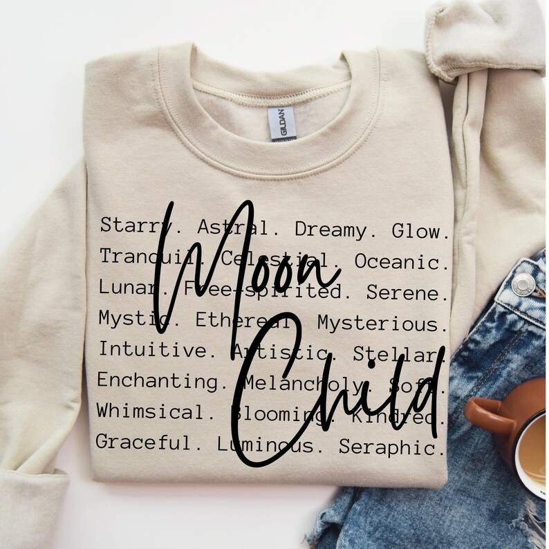 May include: A beige sweatshirt with the words "Moon Child" printed in black. The words are surrounded by a list of adjectives describing the moon, such as "starry", "astral", "dreamy", "glow", "tranquil", "celestial", "oceanic", "lunar", "free-spirited", "serene", "mystic", "ethereal", "mysterious", "intuitive", "artistic", "stellar", "enchanting", "melancholy", "whimsical", "blooming", "kindred", "graceful", "luminous", and "seraphic".