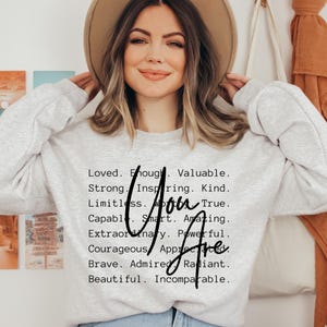 May include: A light gray crewneck sweatshirt featuring black text that reads: Loved. Enough. Valuable. Strong. Inspiring. Kind. Limitless. Worthy. True. Capable. Smart. Amazing. Extraordinary. Powerful. Courageous. Appreciated. Brave. Admired. Radiant. Beautiful. Incomparable.