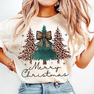 Könnte beinhalten: Ein cremefarbenes T-Shirt mit einer Grafik von drei Weihnachtsbäumen. Die beiden äußeren Bäume haben ein braun-schwarzes Leopardenmuster, während der mittlere Baum einfarbig grün ist und eine große Leoparden-Schleife trägt. Darunter steht Merry Christmas.