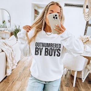 May include: White sweatshirt with the text "OUTNUMBERED BY BOYS" in a bold, black font. The long-sleeved crewneck top is made of a soft material. The sweatshirt is a casual style.