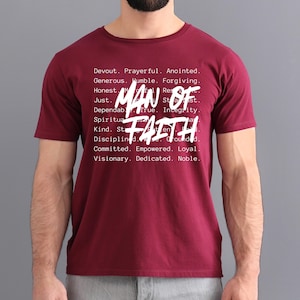 Op de afbeelding: Een bordeauxrood t-shirt met de woorden "MAN OF FAITH" in grote witte letters. Het shirt heeft ook een lijst met positieve eigenschappen in kleinere witte letters, waaronder "Devout", "Prayerful" en "Anointed".