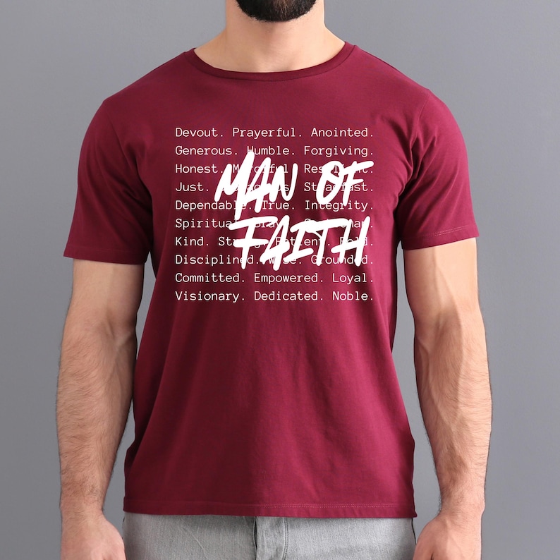 Op de afbeelding: Een bordeauxrood t-shirt met de woorden "MAN OF FAITH" in grote witte letters. Het shirt heeft ook een lijst met positieve eigenschappen in kleinere witte letters, waaronder "Devout", "Prayerful" en "Anointed".