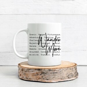 K&ouml;nnte beinhalten: Wei&szlig;e Keramiktasse mit den Worten "Wander More" in handschriftlicher Schrift. Die Tasse ist mit W&ouml;rtern wie "Scenic", "Adventurous" und "Joyful" verziert. Die Tasse steht auf einem runden Holzuntersetzer.