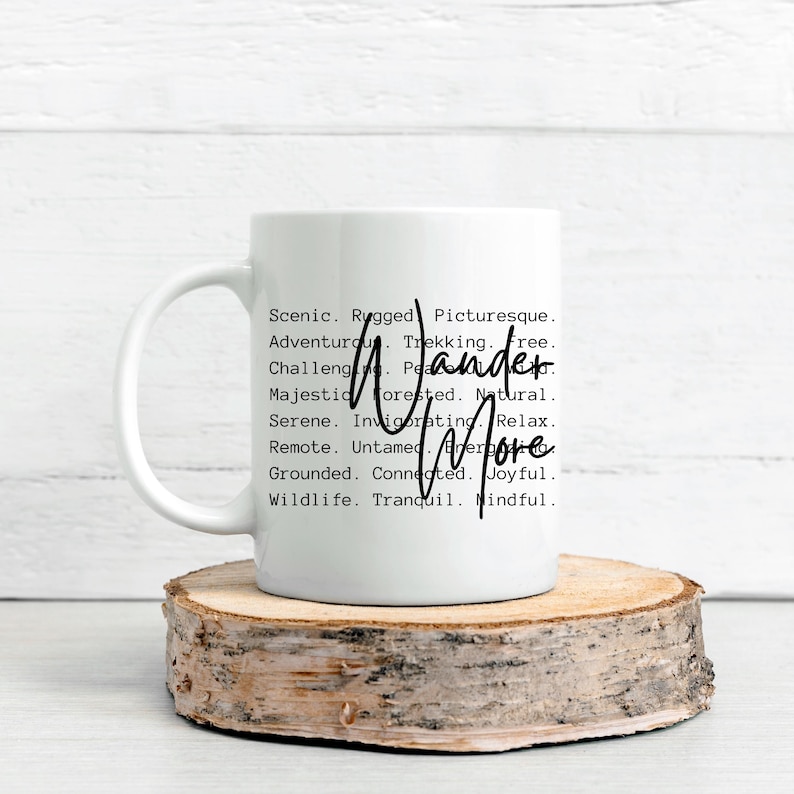 K&ouml;nnte beinhalten: Wei&szlig;e Keramiktasse mit den Worten "Wander More" in handschriftlicher Schrift. Die Tasse ist mit W&ouml;rtern wie "Scenic", "Adventurous" und "Joyful" verziert. Die Tasse steht auf einem runden Holzuntersetzer.