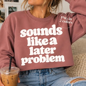 Puede incluir: Una sudadera de cuello redondo color malva con el texto blanco desgastado "sounds like a later problem" impreso en el pecho. La prenda tiene mangas largas y un dobladillo corto. Un gráfico circular en la esquina superior derecha dice "PNG in 2 colors".