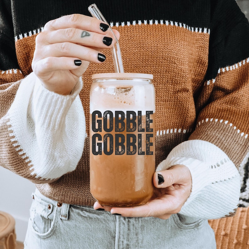 May include: A clear glass beverage can-shaped cup with a light brown drink and a glass straw. The cup has a light pink lid and the words "GOBBLE GOBBLE" in black block letters. The person is wearing a brown, black, and white sweater.