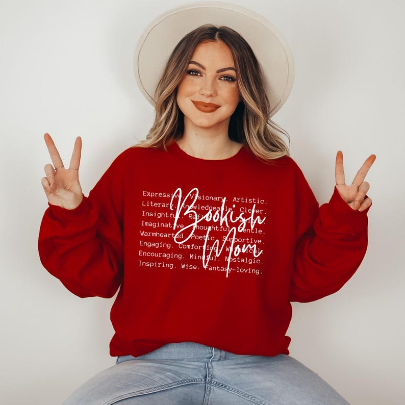 Puede incluir: Sudadera roja con texto blanco que dice "Bookish Mom" y una lista de palabras que describen las cualidades de una madre que le gusta leer.