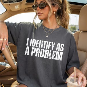 May include: A charcoal gray short-sleeve t-shirt featuring the white distressed-style text "I IDENTIFY AS A PROBLEM" printed across the chest. The shirt has a crew neckline and a relaxed fit, worn with gold-toned hoop earrings and a layered necklace.
