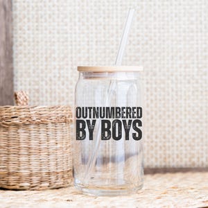 May include: A clear glass can-shaped tumbler with a bamboo lid and a clear straw. The text "OUTNUMBERED BY BOYS" is printed in black on the front. The tumbler is on a neutral background with a woven basket.