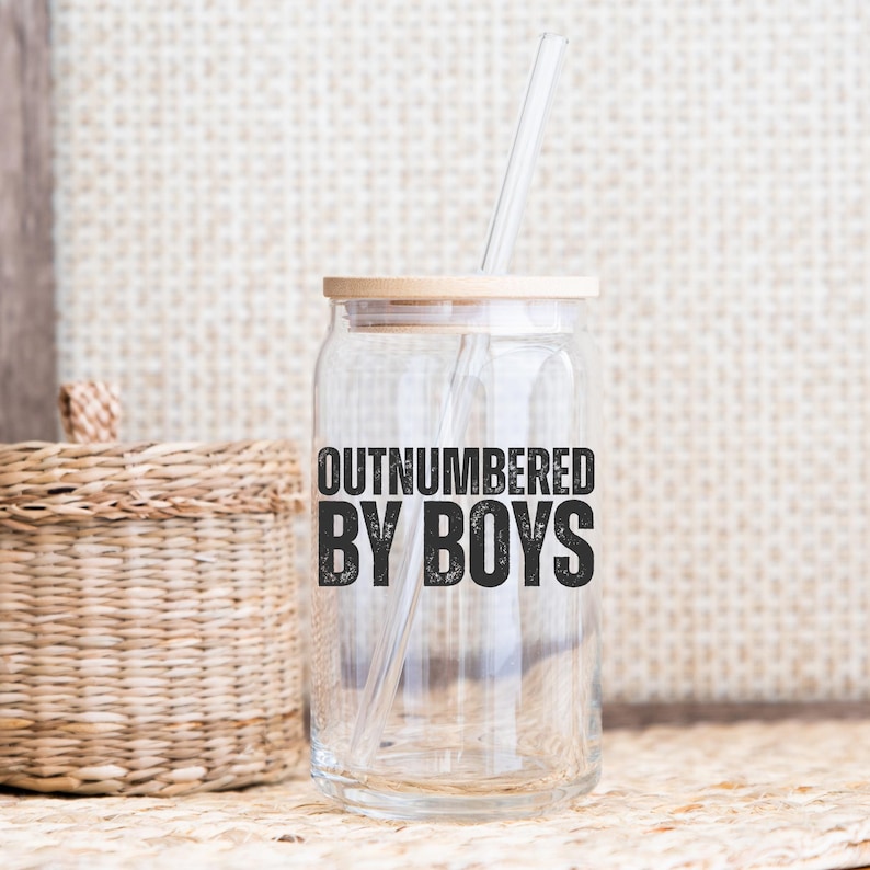 May include: A clear glass can-shaped tumbler with a bamboo lid and a clear straw. The text "OUTNUMBERED BY BOYS" is printed in black on the front. The tumbler is on a neutral background with a woven basket.