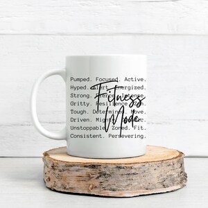 May include: White ceramic mug with the words "Fitness Mode" in a handwritten font. The mug also features a list of motivational words like "Pumped," "Focused," and "Strong." The mug is placed on a wooden slice.