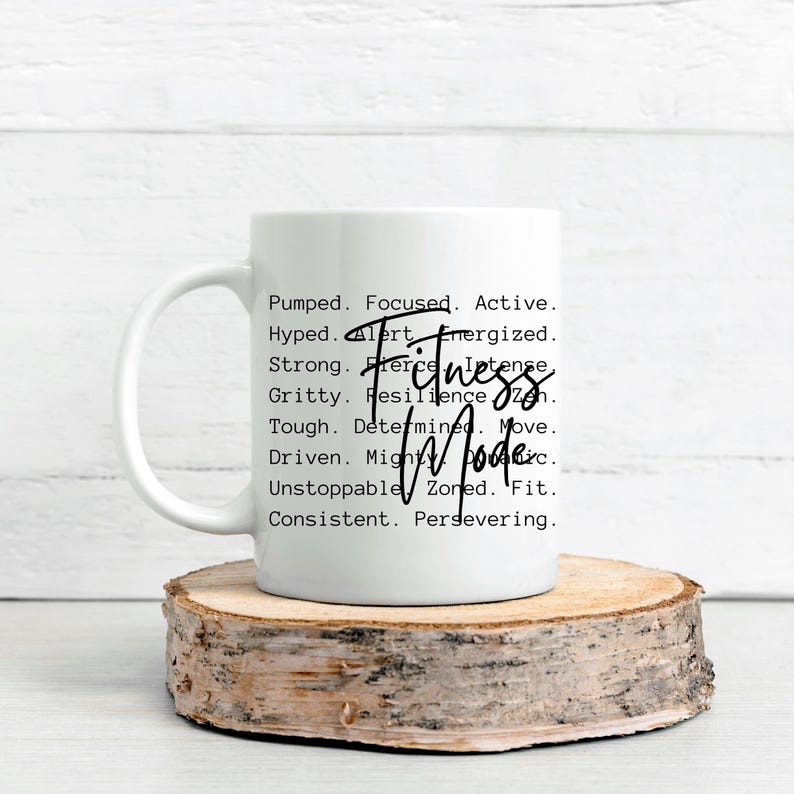 May include: White ceramic mug with the words "Fitness Mode" in a handwritten font. The mug also features a list of motivational words like "Pumped," "Focused," and "Strong." The mug is placed on a wooden slice.