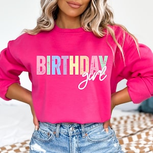 Może przedstawiać: Jasnor&oacute;żowa bluza z napisem "BIRTHDAY girl". Słowo "BIRTHDAY" jest w stylu blokowym z pastelowymi kolorami, a "girl" jest w kursywie. Modelka ma na sobie niebieskie dżinsowe szorty.
