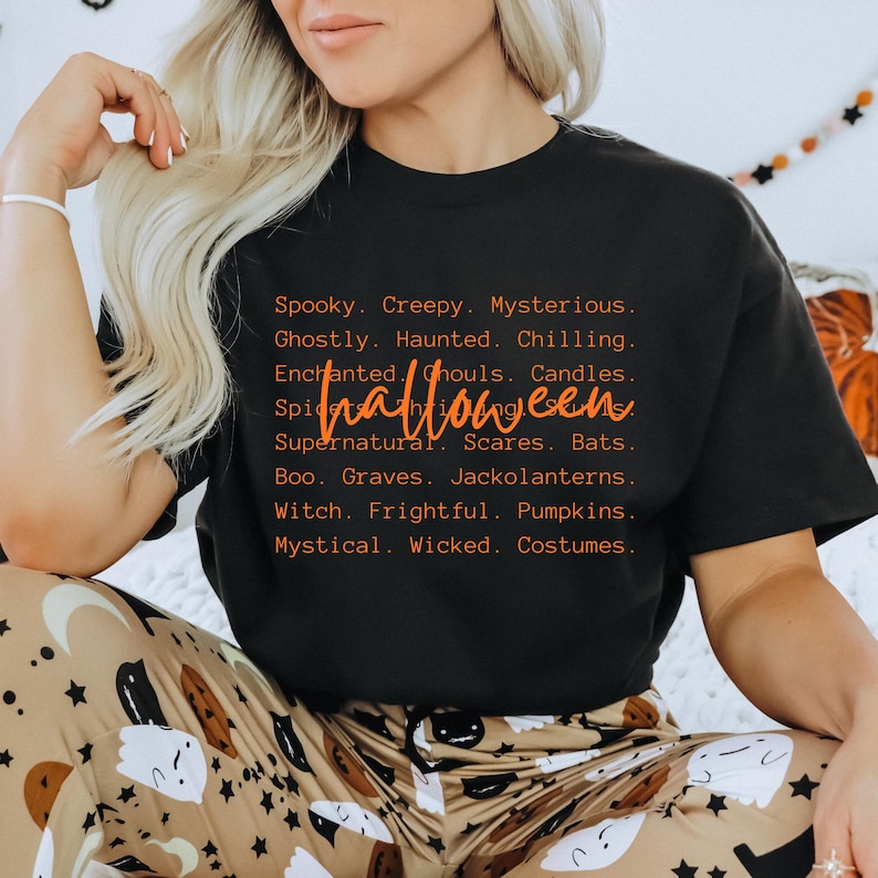 May include: Black t-shirt with orange text that reads "Halloween" and a list of spooky words. The shirt is part of a Halloween-themed collection. The model is wearing patterned pajama pants.