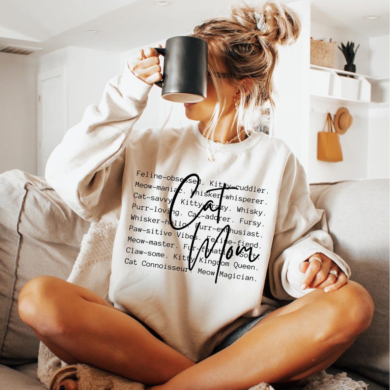 May include: A cream-colored sweatshirt with the words "Cat Mom" in a script font. The sweatshirt has a list of cat-related words in a black font. A person is holding a black mug.