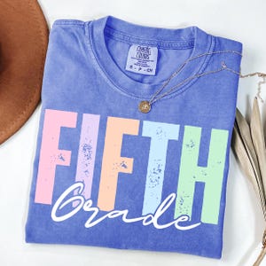 K&ouml;nnte beinhalten: Ein blaues Comfort Colors T-Shirt mit den Worten "FIFTH Grade" in bunten Blockbuchstaben. Eine goldene Halskette mit einem kleinen Anh&auml;nger liegt auf dem Shirt. Das Shirt ist gefaltet und hat einen Vintage-Look.
