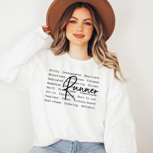 May include: White sweatshirt with a black text graphic that reads "Gritty. Unstoppable. Resilient. Relentless. Zenacious. Focused. Dedicated. Persevering. Driven. Momentum. Runner Swift. Trailblazer. Powerhouse. All-in. Explosive. Born to run. Fearless. Paced. Victory-bound. Goal-chase. Enduring. Athletic."