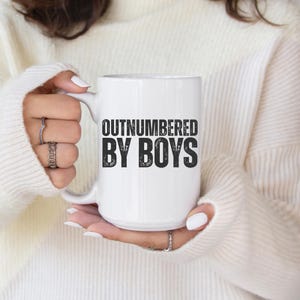 May include: White ceramic mug with the text "OUTNUMBERED BY BOYS" in black block letters. The mug is held by a person wearing a white sweater. The mug has a white handle.