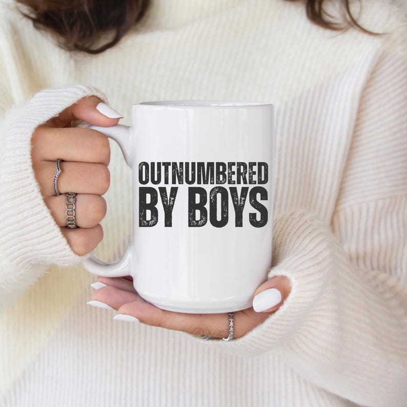 May include: White ceramic mug with the text "OUTNUMBERED BY BOYS" in black block letters. The mug is held by a person wearing a white sweater. The mug has a white handle.