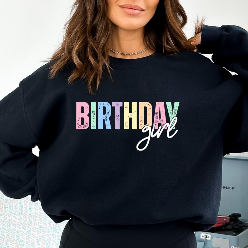 Może przedstawiać: Czarna bluza z napisem "BIRTHDAY girl" w pastelowych literach. Słowo "BIRTHDAY" jest w bloku, a "girl" jest kursywą. Bluza ma okrągły dekolt.