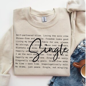 May include: A beige sweatshirt with a black text graphic that reads "Self-partnered bliss. Loving the solo vibe. Stress-free and single. Freedom looks good. Living my best life. Table for one, please. No strings, no stress. Peace over partners. Blankets all mine. Unattached and loving it. Happily unbothered. Single. I am the main character. Time spent for me. Happily my own plus-one. Single, not sorry. Diagonally sleeping goals. Drama-free zone. Me time = best time. Unapologetically solo. No drama, just peace. Single, not mingling."