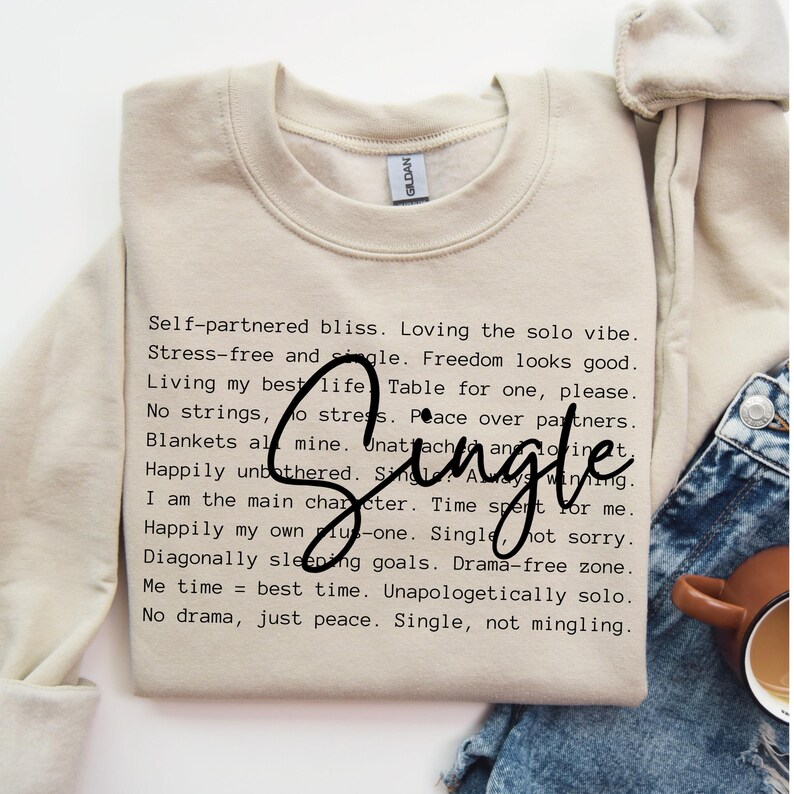 May include: A beige sweatshirt with a black text graphic that reads "Self-partnered bliss. Loving the solo vibe. Stress-free and single. Freedom looks good. Living my best life. Table for one, please. No strings, no stress. Peace over partners. Blankets all mine. Unattached and loving it. Happily unbothered. Single. I am the main character. Time spent for me. Happily my own plus-one. Single, not sorry. Diagonally sleeping goals. Drama-free zone. Me time = best time. Unapologetically solo. No drama, just peace. Single, not mingling."