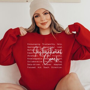 May include: A red sweatshirt with the words "Instructional Coach" in white script. The sweatshirt also features a list of positive attributes in white text. The person in the image is wearing a beige beanie.