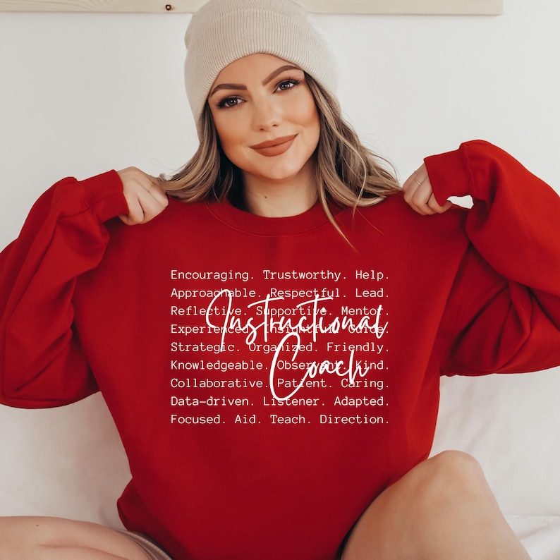 May include: A red sweatshirt with the words "Instructional Coach" in white script. The sweatshirt also features a list of positive attributes in white text. The person in the image is wearing a beige beanie.