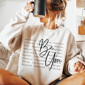 May include: A cream-colored crewneck sweatshirt featuring black text that lists various positive adjectives including Authentic, Expressive, Fearless, Original, Honest, True, Genuine, Bold, Unapologetic, Independent, Natural, Brave, Confident, Unfiltered, Unique, Real, Whole, Radiant, Individual, Courageous, Creative, Unmasked, Unbreakable, Strong, Limitless, Open, Bright, Balanced, Empowered, Determined, with the words Be You written in a large black script font over the text.