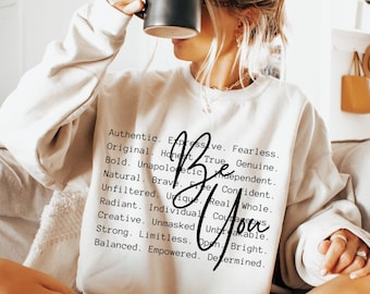 May include: A cream-colored crewneck sweatshirt featuring black text that lists various positive adjectives including Authentic, Expressive, Fearless, Original, Honest, True, Genuine, Bold, Unapologetic, Independent, Natural, Brave, Confident, Unfiltered, Unique, Real, Whole, Radiant, Individual, Courageous, Creative, Unmasked, Unbreakable, Strong, Limitless, Open, Bright, Balanced, Empowered, Determined, with the words Be You written in a large black script font over the text.