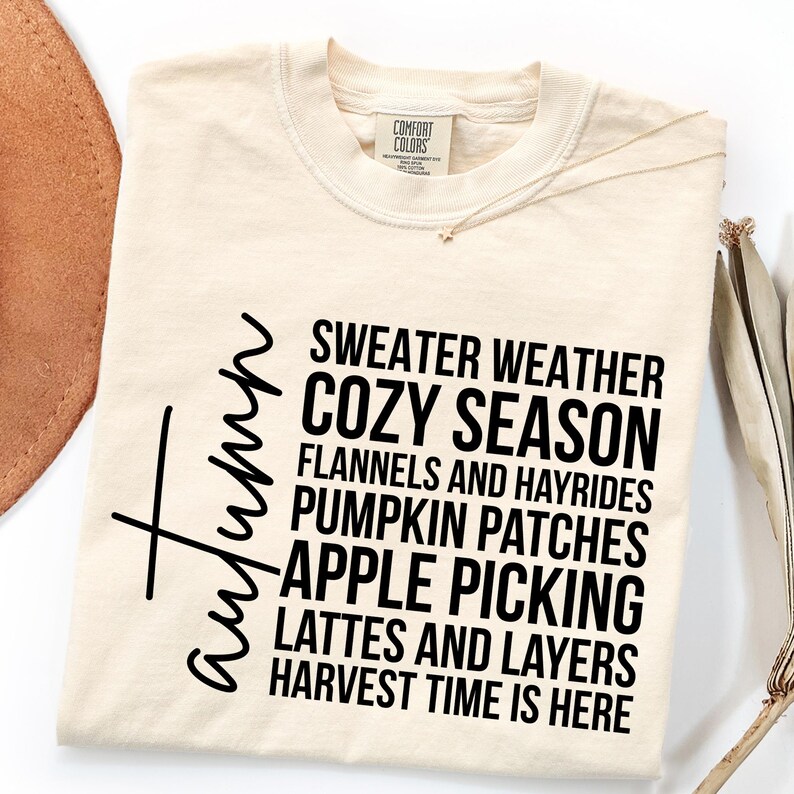K&ouml;nnte beinhalten: Cremefarbenes T-Shirt mit dem Wort "Autumn" in Schreibschrift. Das Shirt zeigt die S&auml;tze "Sweater Weather", "Cozy Season" und "Harvest Time is Here" in fetter, schwarzer Schrift, die den Herbst hervorrufen.
