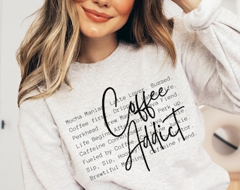 May include: A light gray crewneck sweatshirt featuring a list of coffee-related phrases in black text, including Mocha Maniac, Coffee first, Brew Master, and Caffeine Connoisseur, with the words Coffee Addict written in a large black script.