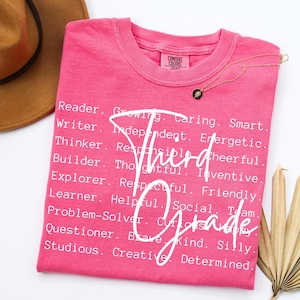 Może przedstawiać: R&oacute;żowa koszulka Comfort Colors z napisem "Third Grade" w kursywie. Koszulka zawiera r&oacute;wnież listę pozytywnych przymiotnik&oacute;w, takich jak "Reader", "Writer" i "Thinker".
