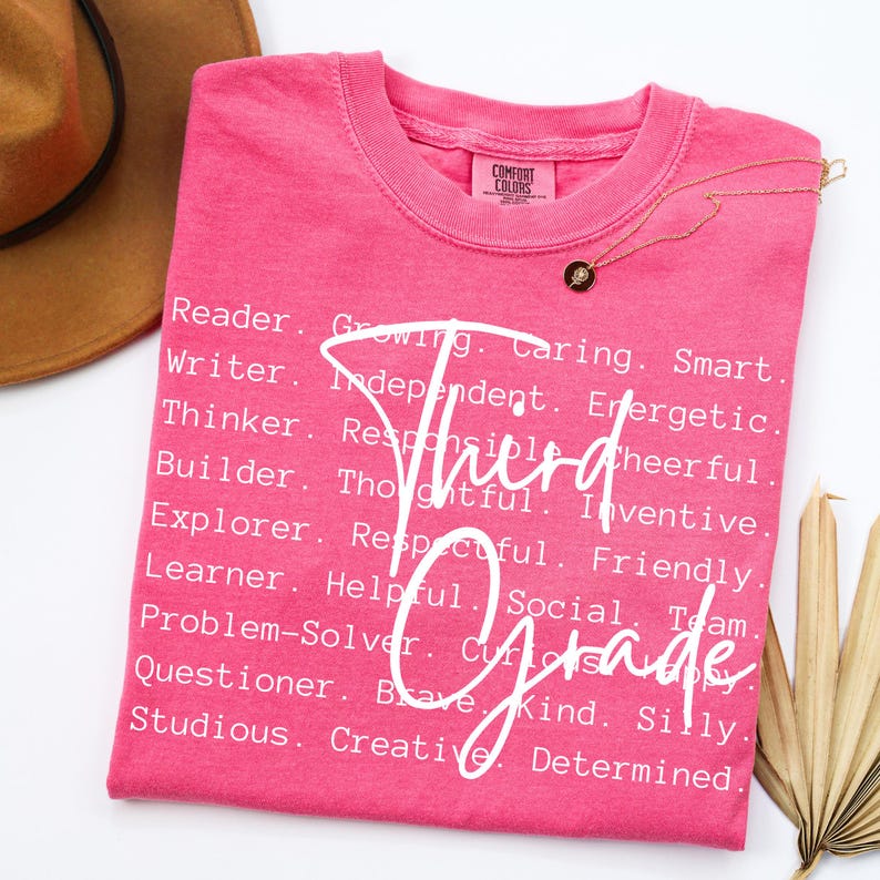 Może przedstawiać: R&oacute;żowa koszulka Comfort Colors z napisem "Third Grade" w kursywie. Koszulka zawiera r&oacute;wnież listę pozytywnych przymiotnik&oacute;w, takich jak "Reader", "Writer" i "Thinker".