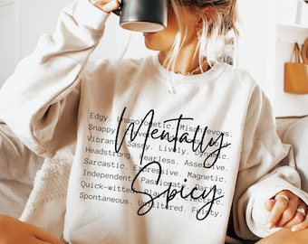 Op de afbeelding: Een crèmekleurige sweater met ronde hals en zwarte tekst Mentally Spicy in een groot cursief lettertype, geplaatst over een lijst met bijvoeglijke naamwoorden zoals Edgy, Unapologetic, Mischievous, Snappy, Clever, Bold, Vibrant, Sassy, Lively en Dynamic.