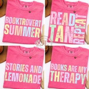 May include: Four pink t-shirts with pastel text. The shirts read "BOOKTROVERT SUMMER", "READ TAN REPEAT", "STORIES AND LEMONADE", and "BOOKS ARE MY THERAPY". Each shirt has a small gold necklace.
