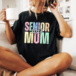 Puede incluir: Camiseta negra con el texto "SENIOR MOM 2027" en colores pastel. La camiseta es de corte holgado y tiene cuello redondo. El texto est&aacute; en una fuente audaz y juguetona, lo que la convierte en un gran regalo.