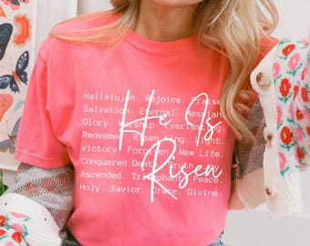 Op de afbeelding: Een fel koraalkleurig T-shirt met korte mouwen en witte tekst He Is Risen, omringd door een lijst met woorden waaronder Hallelujah, Rejoice, Praise, Salvation, Eternal, Messiah, Glory, Worship, Everlasting, Redeemed, Risen King, Light, Victory, Forgiven, New Life, Conquered Death, Truth, Joy, Ascended, Triumphant, Peace, Holy, Savior, Grace en Divine.