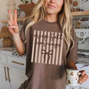 May include: Brown t-shirt with the text "crisp fall mornings" in a vertical striped design. The shirt also features a bow graphic. A white mug with bear illustrations is held in the hand.
