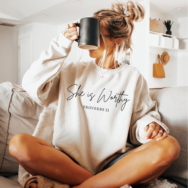 May include: A cream-coloured sweatshirt featuring the script "She is Worthy" and "Proverbs 31" in black. A person is holding a black mug. The sweatshirt is a neutral tone and looks to be made of a soft fabric.