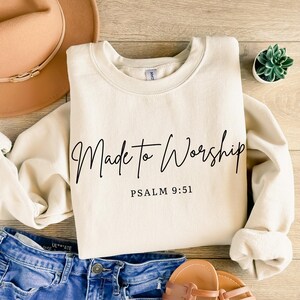 May include: Cream-colored sweatshirt with the words "Made to Worship" in black script, and the text "Psalm 9:51" below. The sweatshirt is on a wooden surface, with a hat, jeans, and sandals in the background.
