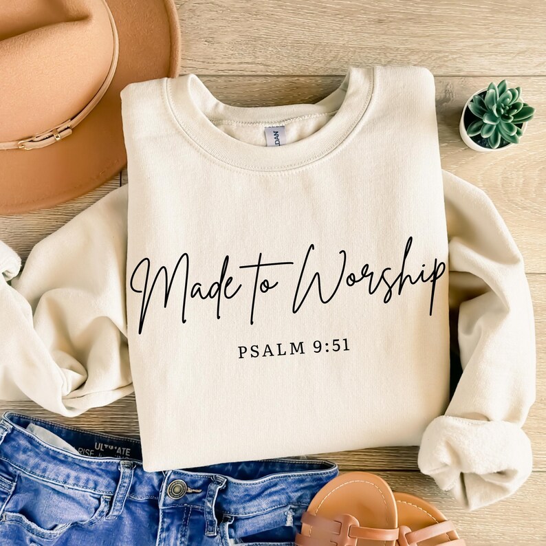 May include: Cream-colored sweatshirt with the words "Made to Worship" in black script, and the text "Psalm 9:51" below. The sweatshirt is on a wooden surface, with a hat, jeans, and sandals in the background.
