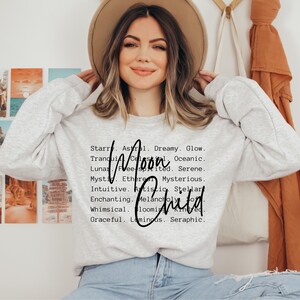 May include: A gray sweatshirt with the words "Moon Child" in black script. The text is surrounded by a list of words describing the moon, such as "dreamy", "glow", "serene", "mysterious", "artistic", and "whimsical".