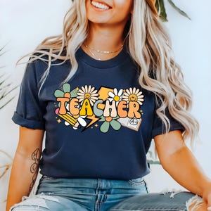 May include: A navy blue t-shirt with a colorful graphic design that says "TEACHER" in a retro font. The graphic includes flowers, a lightning bolt, a pencil, and a book with an A+ grade.