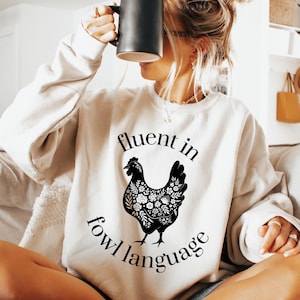 May include: A cream-colored crewneck sweatshirt featuring a black graphic of a chicken filled with floral patterns. The text "fluent in fowl language" is printed in a black serif font, arching above and curving below the chicken illustration.