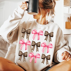 May include: A cream-colored crewneck sweatshirt featuring a repeating pattern of nine bows arranged in a grid. The design alternates between solid pink bows and brown and black leopard print bows, all rendered in a graphic illustration style.