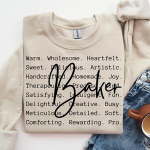 May include: A beige sweatshirt with the text "Warm. Wholesome. Heartfelt. Sweet. Delicious. Artistic. Handcrafted. Homemade. Joy. Therapeutic. Precise Baker Satisfying. Indulgent. Fun. Delightful. Creative. Busy. Meticulous. Detailed. Soft. Comforting. Rewarding. Pro." printed on it.