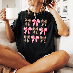 Puede incluir: Una camiseta negra de manga corta con un patr&oacute;n de cuadr&iacute;cula de nueve lazos. El dise&ntilde;o alterna entre lazos de color rosa s&oacute;lido y lazos con estampado de leopardo marr&oacute;n y negro, dispuestos en tres filas de tres en el centro del pecho.